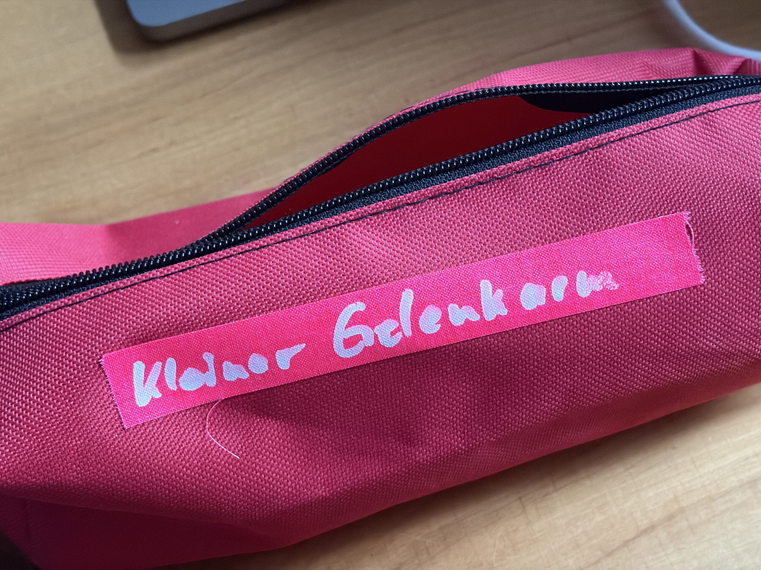 Pinke Reißverschlusstasche mit weißer Beschriftung „Kleiner Gelenkarm“ auf pinkem Klebeband